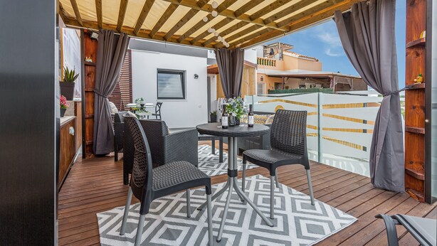 Un patio exterior con terraza de madera, una mesa redonda, dos sillas y una pérgola.