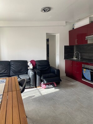 A living room with a sofa, a small table, and a kitchen area with red cabinets and a TV.