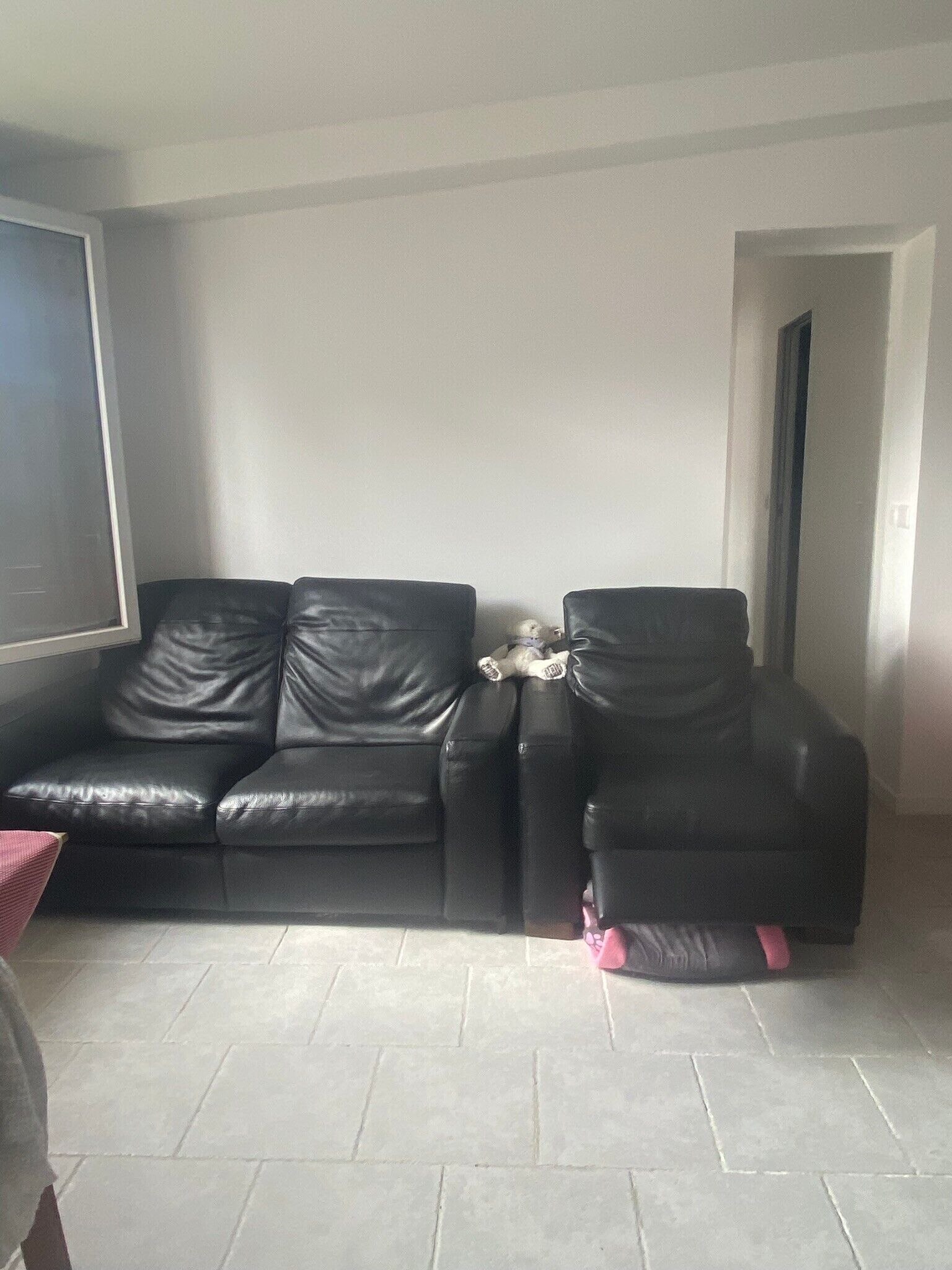 Una habitación con un sofá de cuero negro y un sillón negro, una mesita con un objeto rosa, y un suelo de baldosas.
