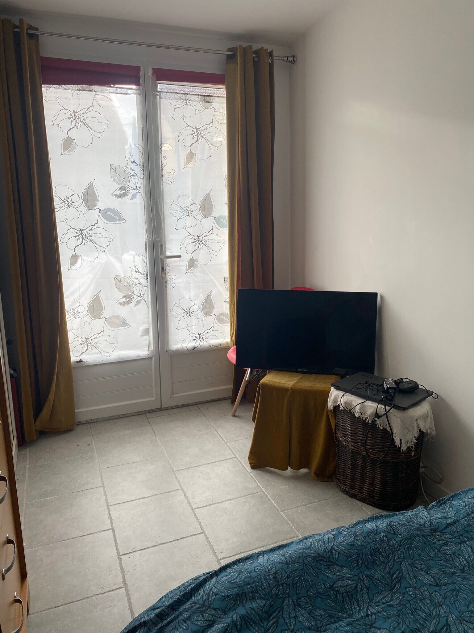 Une pièce avec un lit, un téléviseur posé sur une table, un panier en osier et une fenêtre avec des rideaux.