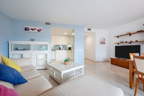 Una sala de estar con sofá, mesa de centro, televisor y cocina al fondo.