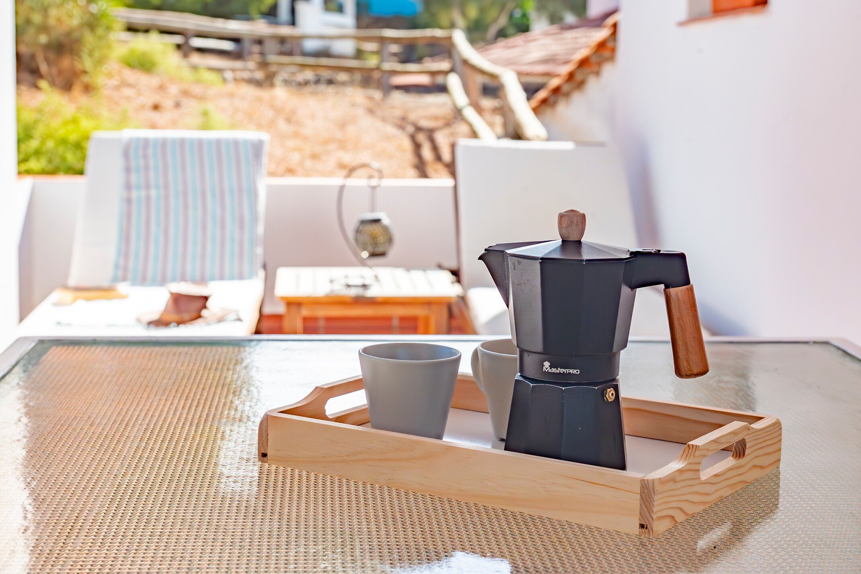 A coffee maker on a tray with two cups, a striped chair, and a table with a view of a garden and building.