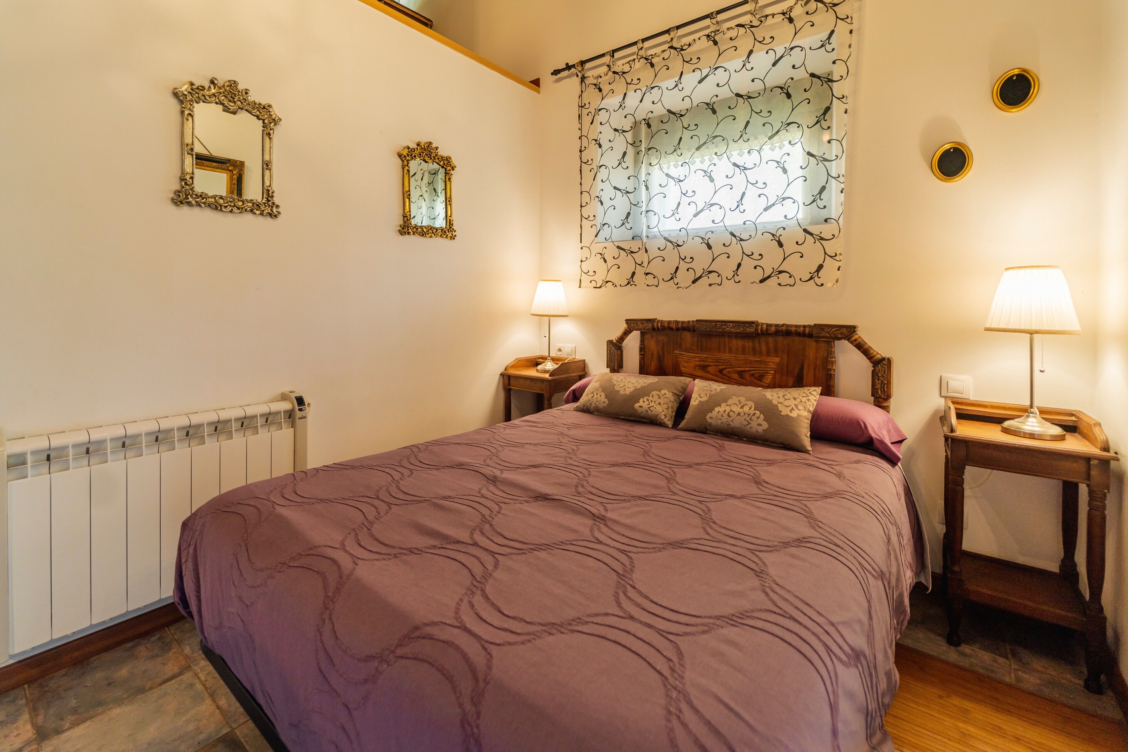 Un dormitorio con una cama grande, dos mesitas de noche, cabecero de madera, un radiador y una ventana con cortinas.