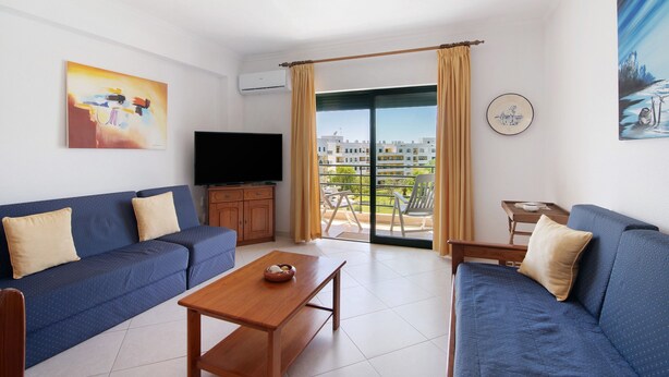A living room with a sofa, a coffee table, a TV, and a balcony with a view of buildings. - Apartment 'Vila Armação de Pêra' with Shared Pool, Wi-Fi and Air Conditioning (Armação de Pêra)