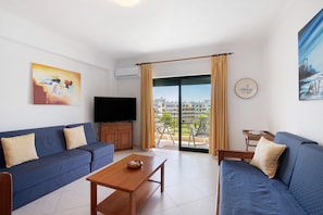 Un salon comprenant un canapé, une table basse, un téléviseur et un balcon offrant une vue sur les immeubles.