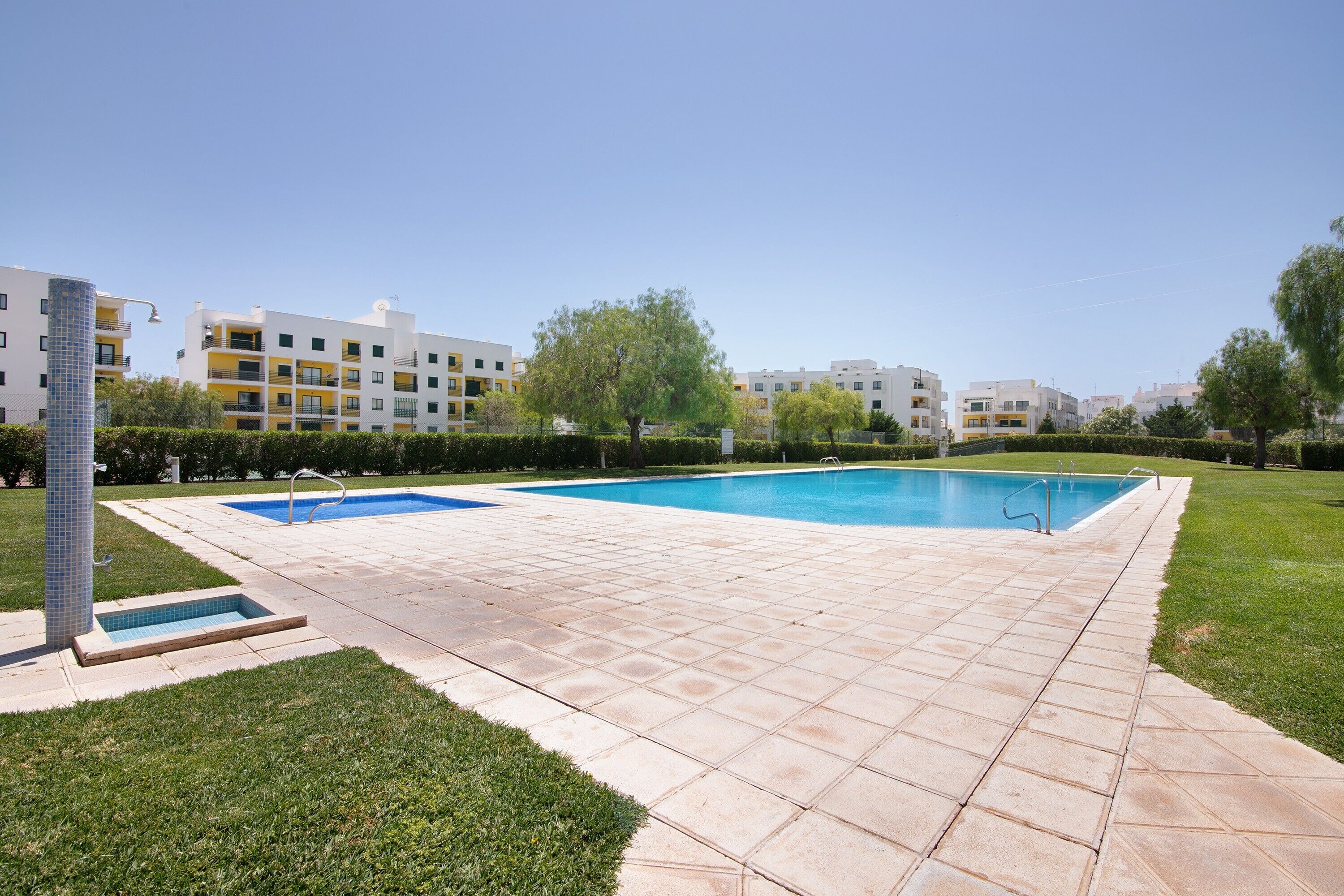 Une piscine dotée d’une terrasse carrelée, entourée d’une pelouse et avec des bâtiments en arrière-plan.