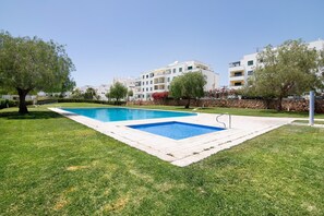 Une piscine rectangulaire dotée d’un plongeoir, entourée d’une pelouse bien entretenue et d’arbres, avec des immeubles à plusieurs étages en arrière-plan.