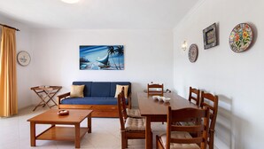 A living room with a sofa, coffee table, dining table, chairs, and a painting of a sailboat. - Apartment 'Vila Armação de Pêra' with Shared Pool, Wi-Fi and Air Conditioning (Armação de Pêra)