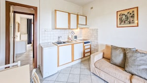 A compact kitchen with a built-in oven, a sink, and a countertop. There is a beige sofa and a framed picture on the wall.