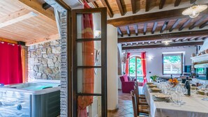 A spacious room with a stone wall, a hot tub, a dining area with a table set for a meal, and a view of the outdoors through a window.