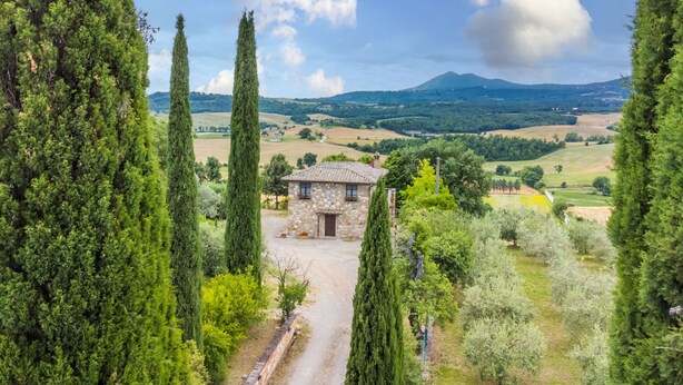 A stone building with a tiled roof surrounded by tall cypress trees and a rural landscape with fields and hills in the background.