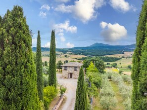 A stone building with a tiled roof surrounded by tall cypress trees and a rural landscape with fields and hills in the background.