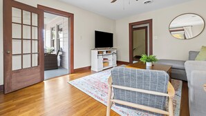 A living room with hardwood flooring, a ceiling fan, a television on a white cabinet, a grey sofa, and a patterned rug.