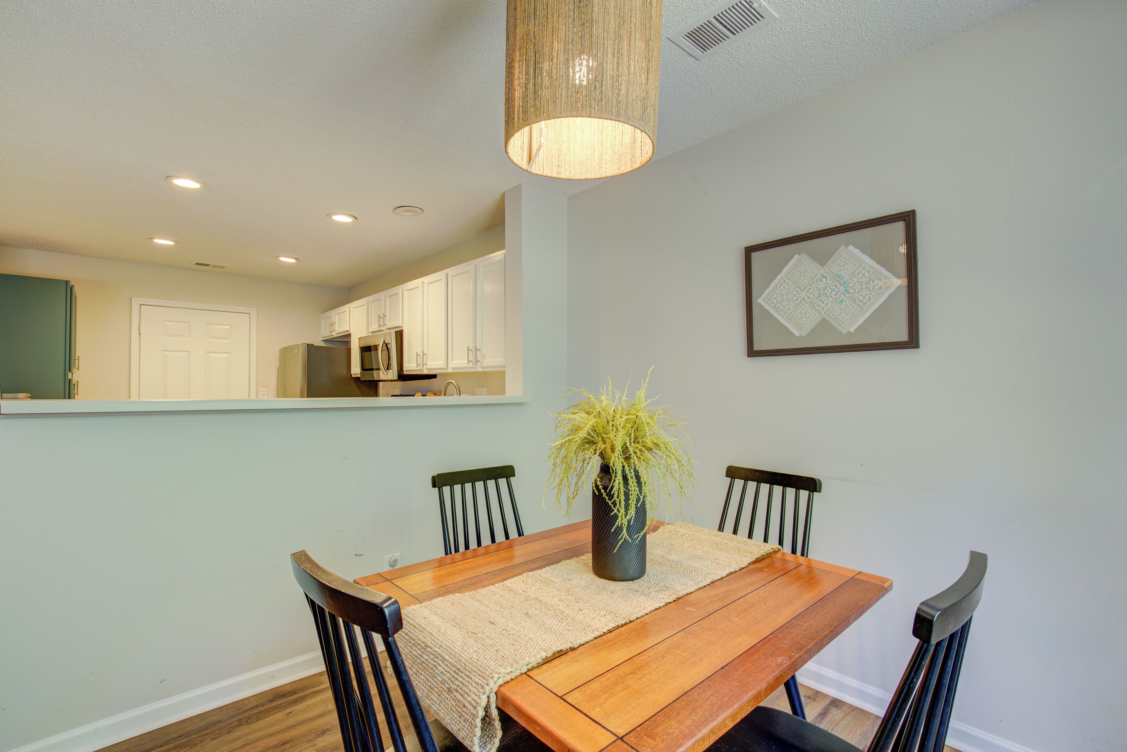 A dining area with a wooden table, four chairs, a vase with a plant, a hanging light fixture, and a kitchen visible in the background.