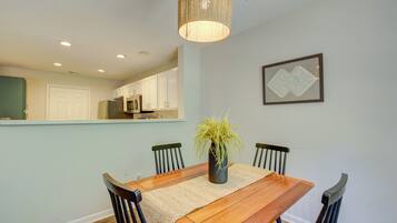 A dining area with a wooden table, four chairs, a vase with a plant, a hanging light fixture, and a kitchen visible in the background.