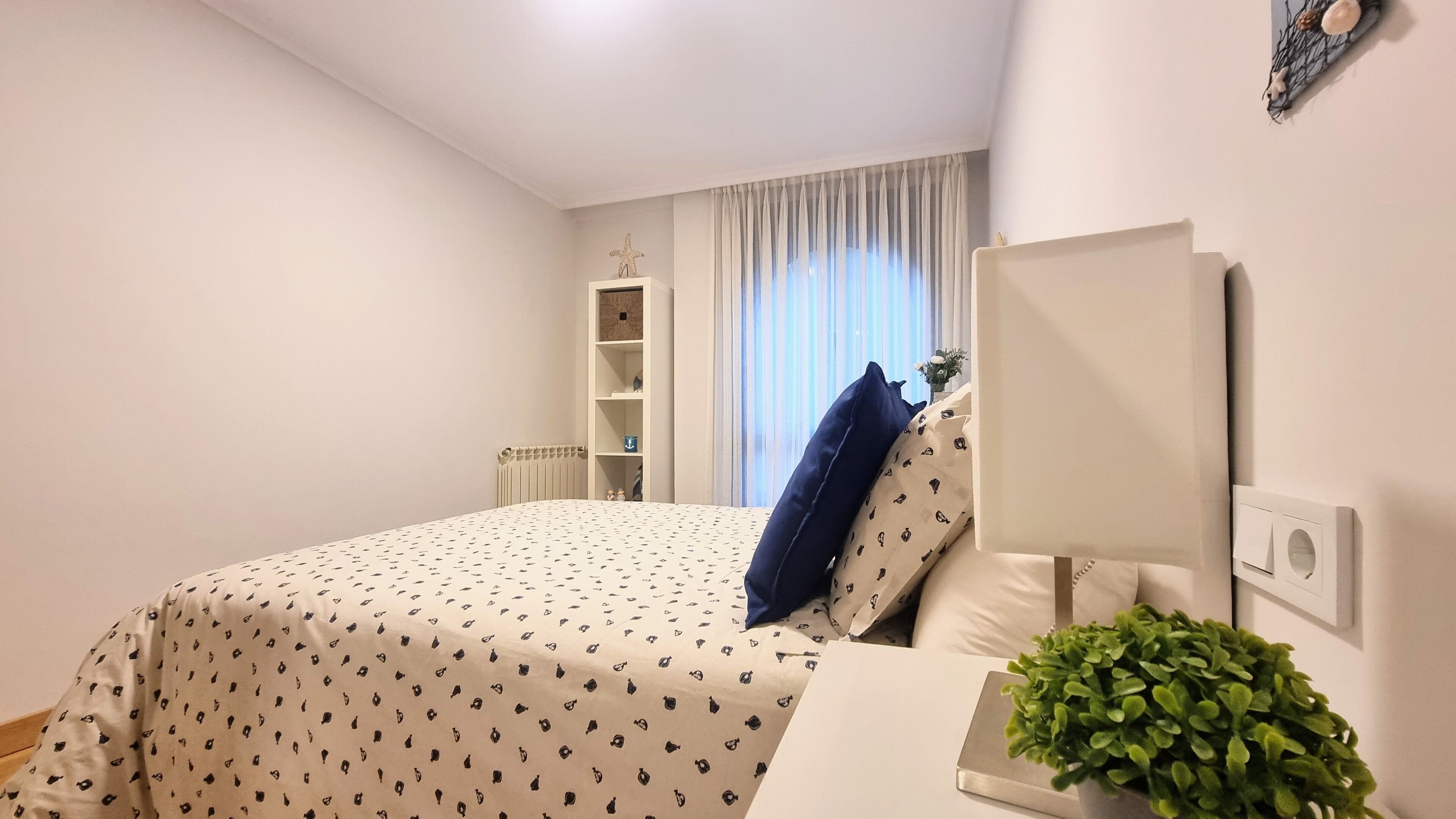 Un dormitorio con una cama, una mesita de noche con lámpara, una planta pequeña y una ventana con cortinas.