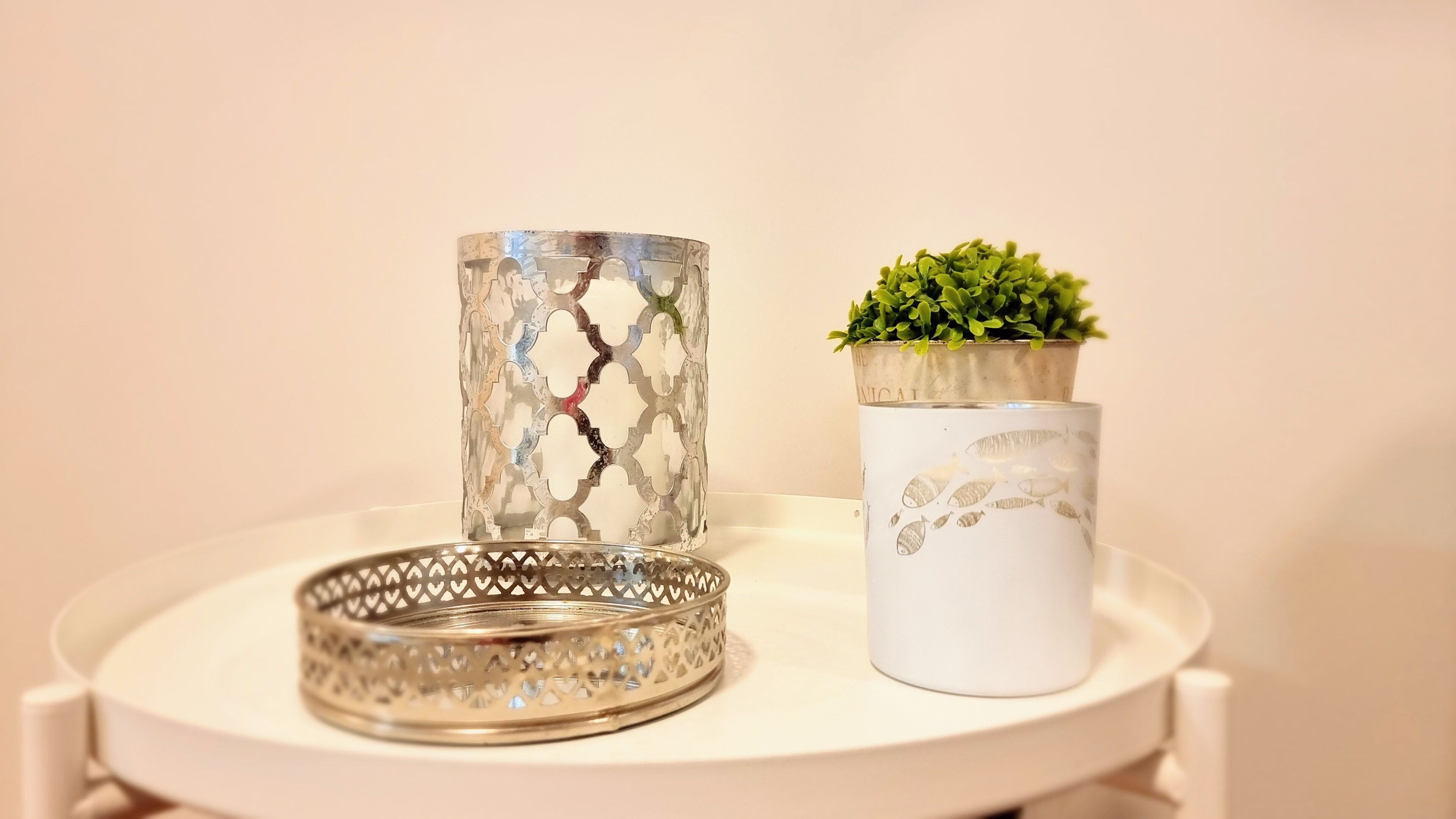 Una mesa blanca con dos adornos: un florero metálico con un diseño agrietado y un florero blanco con motivos de peces, además de una pequeña planta verde en una maceta.
