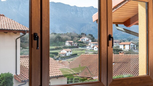 A view through a wooden-framed window of a residential area with terracotta-tiled roofs, surrounded by mountains. - Villa Sonrisa de Llanes (Pancar)