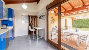 A modern kitchen with a blue cabinet, a white countertop, and a stainless steel refrigerator. There is a dining area with a white table and chairs, and a large window with a view of a green hedge. - Villa Sonrisa de Llanes (Pancar)