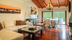 A living room with a sofa, coffee table, and dining area with chairs and a table. There is a painting on the wall, a chandelier, and a large window with a view of greenery. - Villa Sonrisa de Llanes (Pancar)