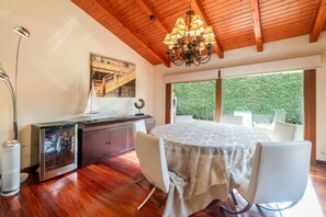 Un espace repas doté d’un plafond en bois, d’un lustre, d’une table à manger avec des chaises, d’un meuble orné d’un objet décoratif et d’une grande fenêtre donnant sur un paysage verdoyant.