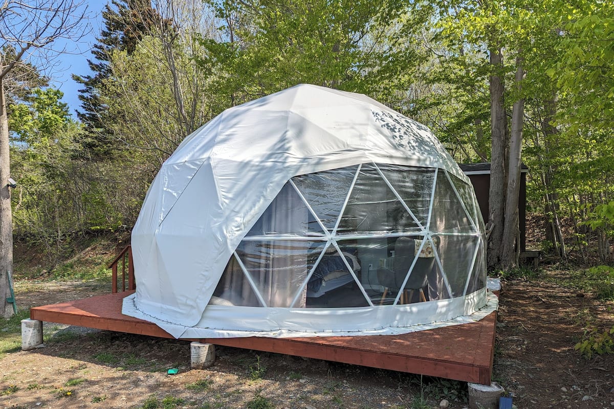 A geodesic dome tent with a transparent window section, set on a wooden platform in a wooded area.