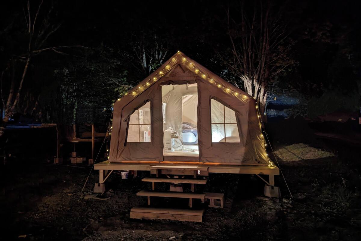 A small, lighted tent with a window and a door, surrounded by trees.