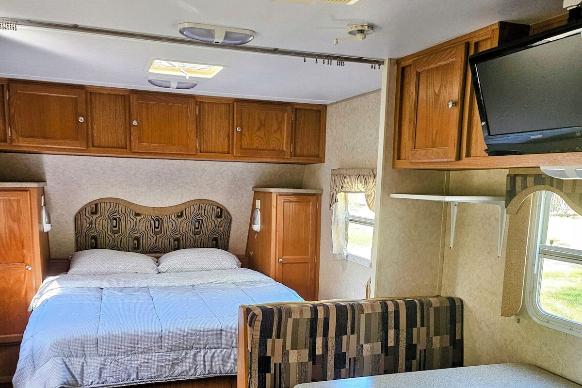 A compact RV interior with a bed, a dining table, and a TV mounted on the wall.