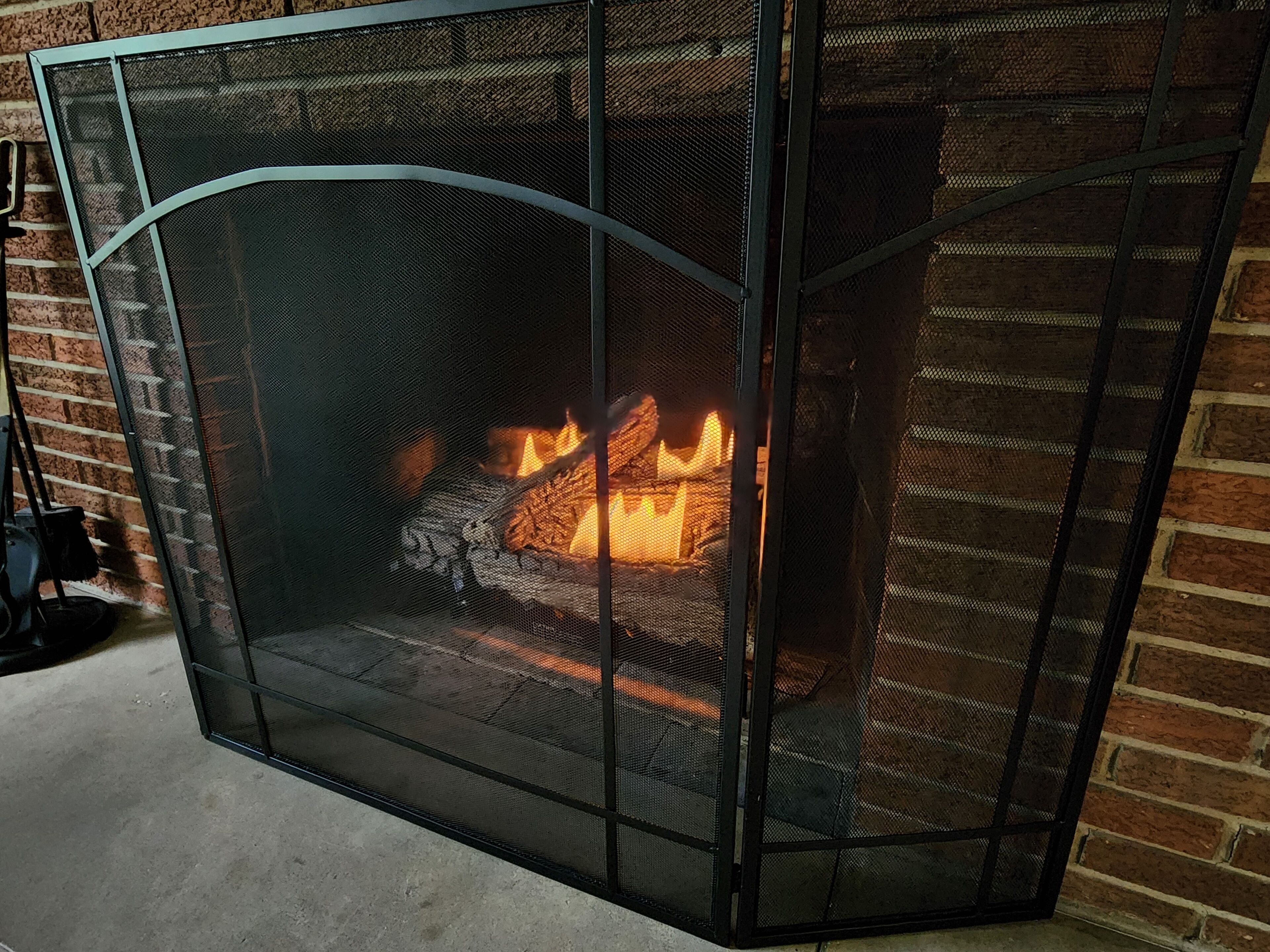 A fireplace with a metal screen, burning logs, and a brick wall in the background.