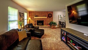 A living room with a brick fireplace, leather furniture, a flat-screen TV, and a bookshelf. - Arcade + Hot Tub Retreat Sauna Near WPAFB (Dayton)