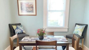 En spisestue med et rektangulært bord, seks stole, en vase med blomster, et indrammet billede på væggen og et vindue med persienner.