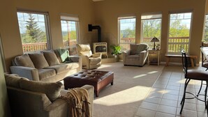 A spacious living room with a beige sofa, a brown ottoman, and a dining area with a bench and chairs. There is a fireplace and a large window with a view of trees. - Amazing view, perfect for large families, central Kenai Peninsula location! (Soldotna)