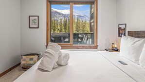 A hotel room with a large window offering a mountain view, a neatly made bed, a bedside table with a lamp, and a framed picture on the wall. - Spring Creek Vacations 1 BR Condo 108TM (Canmore)