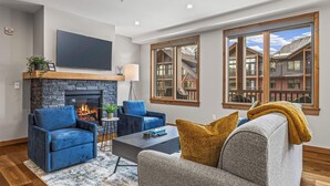 A living room with a fireplace, blue and gray sofas, a coffee table, and large windows with a view of a mountain cabin. - Spring Creek Vacations 2 BR Premier Condo 315TM (Canmore)
