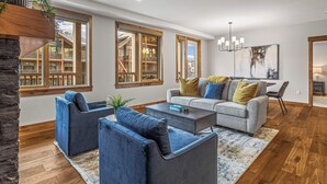 A modern living room with a grey sofa, blue armchairs, and a coffee table. There are large windows with wooden frames, a chandelier, and a painting on the wall. - Spring Creek Vacations 2 BR Premier Condo 315TM (Canmore)