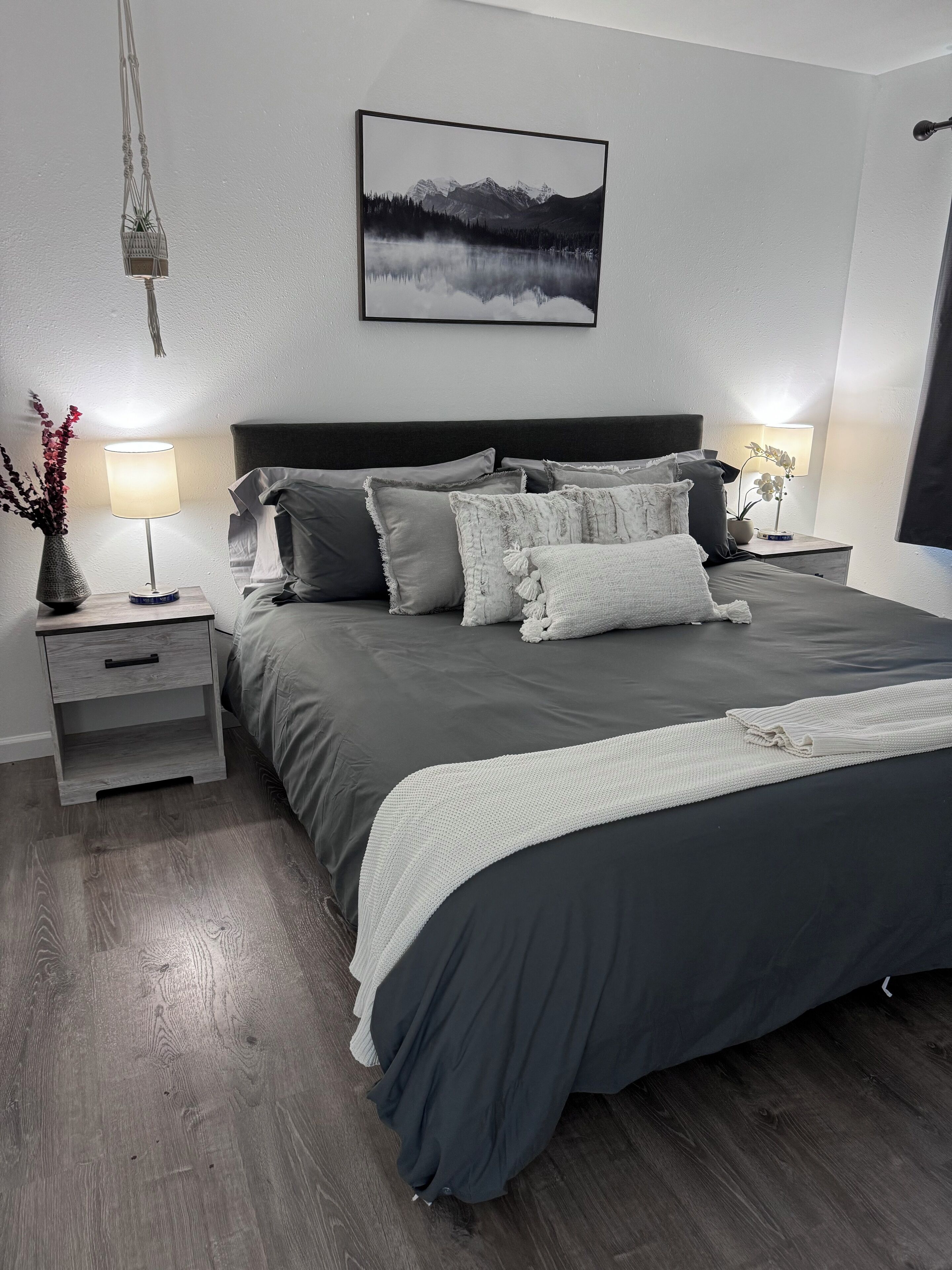 A modern bedroom with a large bed, two bedside tables, a nightstand, a wall-mounted picture, and a hanging macrame piece.