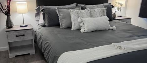 A modern bedroom with a large bed, two bedside tables, a nightstand, a wall-mounted picture, and a hanging macrame piece.