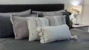Una cama bien hecha con ropa de cama gris y varios almohadones, una mesita de noche con una lámpara y una pequeña planta en maceta, y una pared blanca al fondo.