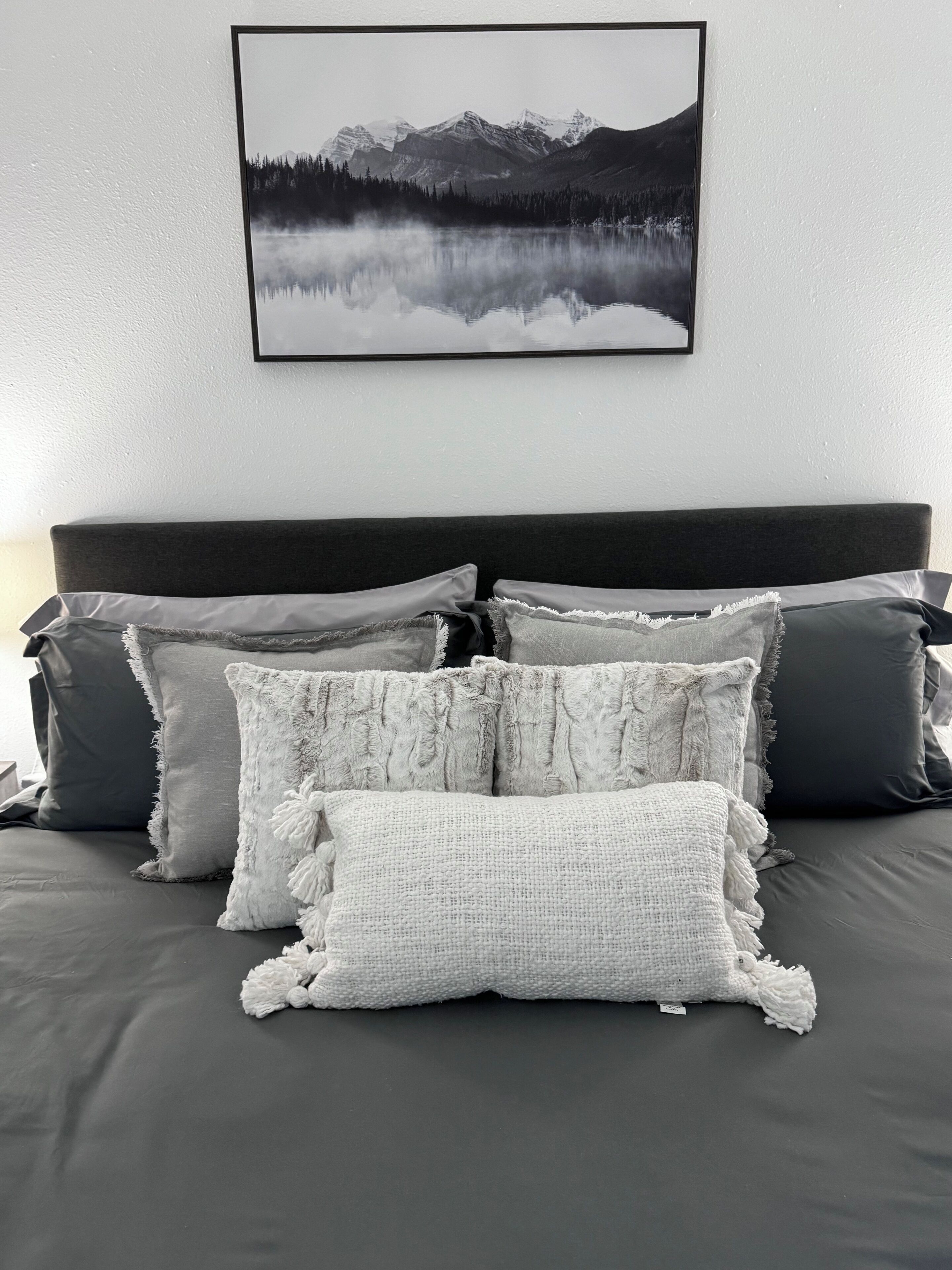 Una cama con ropa de cama gris y múltiples almohadas, incluyendo una almohada blanca con textura.