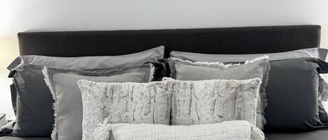Una cama con ropa de cama gris y múltiples almohadas, incluyendo una almohada blanca con textura.