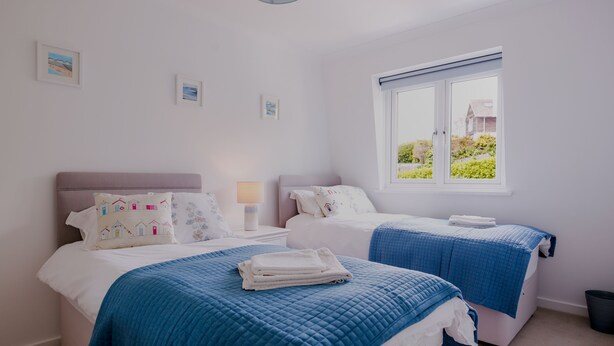 Une chambre avec deux lits, chacun recouvert d’une couette bleue, avec du linge de lit blanc et des oreillers assortis. On peut voir des tableaux encadrés au mur, une fenêtre donnant sur un paysage verdoyant et une table de chevet avec une lampe.