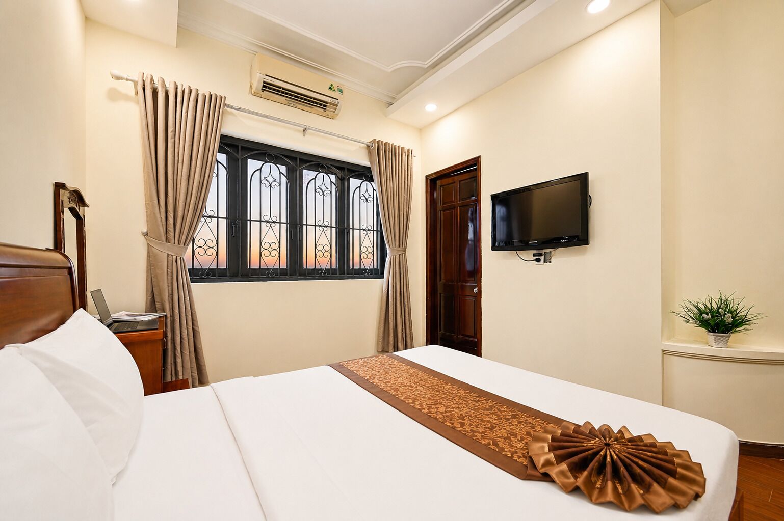 A hotel room with a bed, a television, a window with curtains, an air conditioning unit, and a small potted plant.