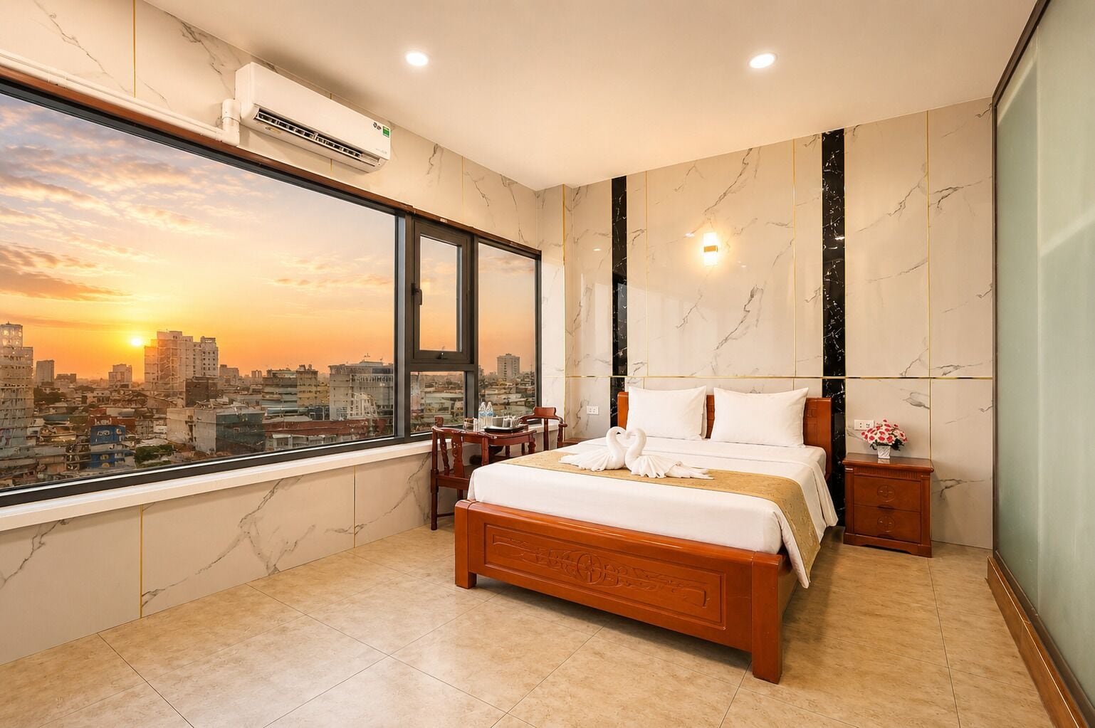 A hotel room with a large bed, a bedside table, a nightstand, a window with a city view, and an air conditioning unit.