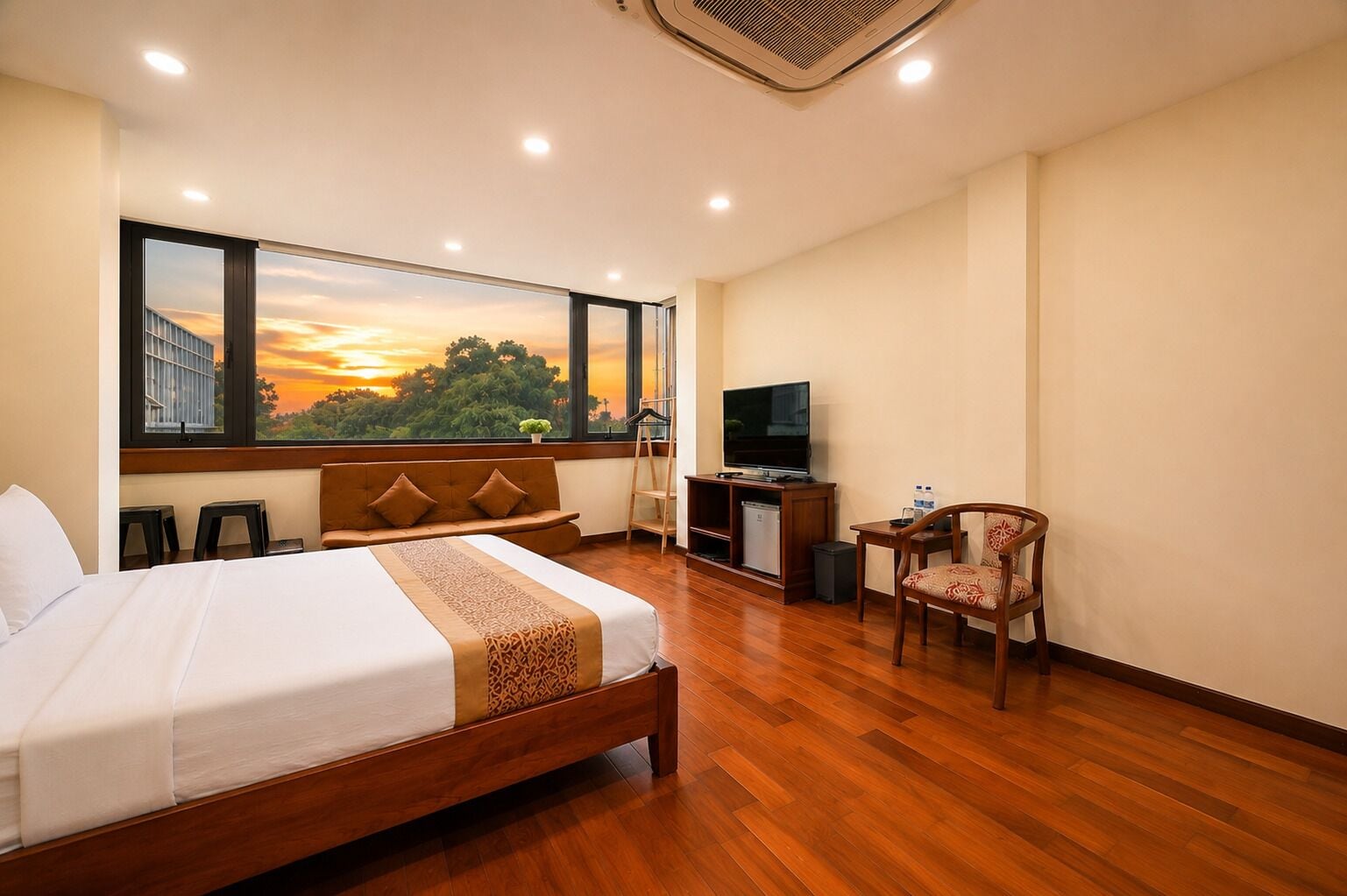 A hotel room with a bed, a TV, a chair, and a view of a sunset through large windows.