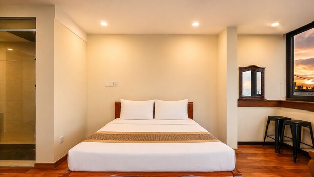 A hotel room with a bed, wooden floor, a window with a sunset view, and a small bar area.