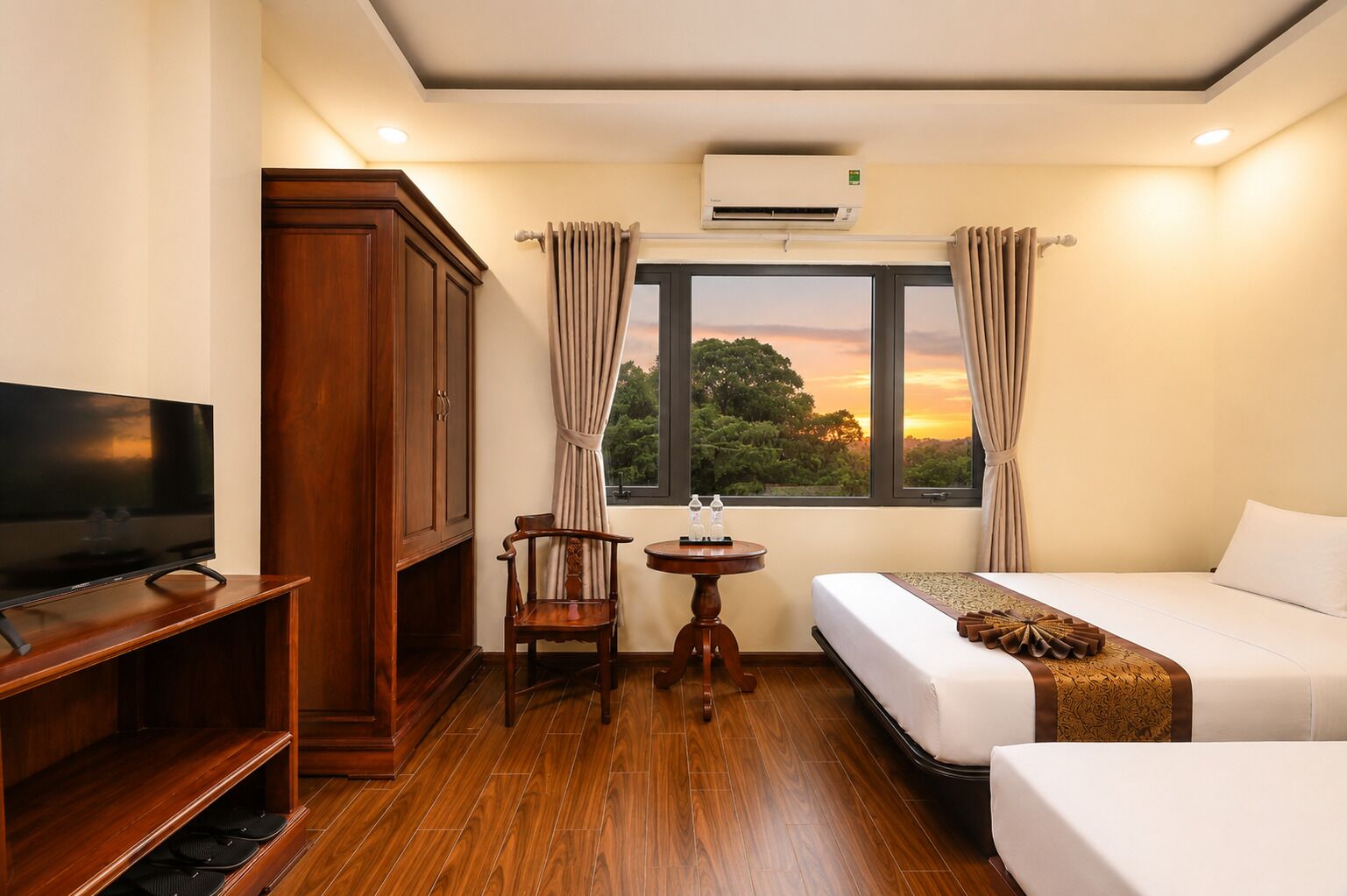 A hotel room with a television, a wooden cabinet, a small table with water bottles, a bed, and a window with curtains.