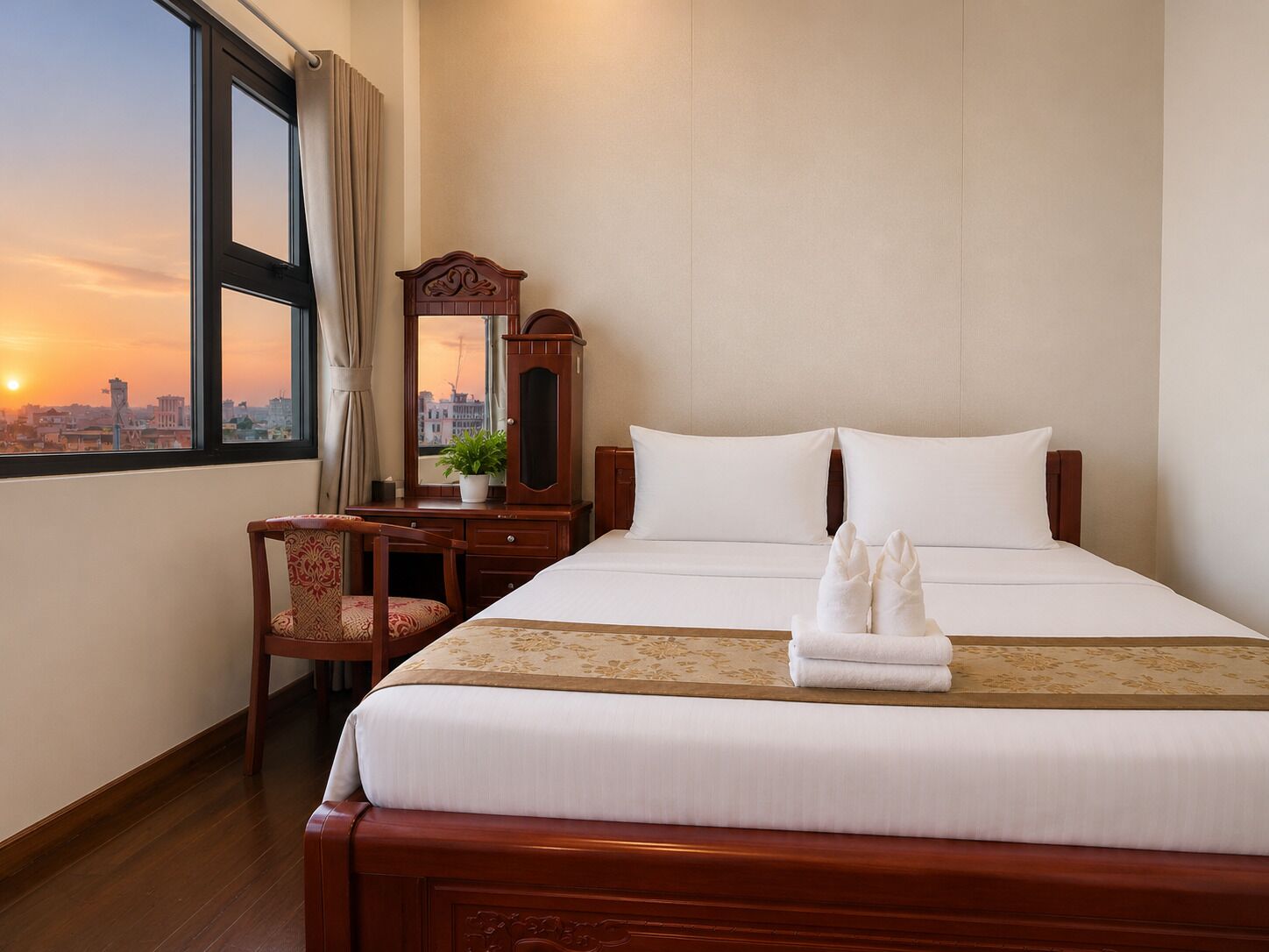 Habitación de hotel con cama, escritorio, silla, espejo y vistas a la ciudad al atardecer.
