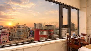 A room with a large window overlooking a cityscape at sunset, a bed, a table with chairs, and a water bottle on the table.