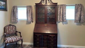 Ein Zimmer mit einem Holzschrank mit Glastüren, einem blumengemusterten Sessel und zwei Fenstern mit floralen Vorhängen.