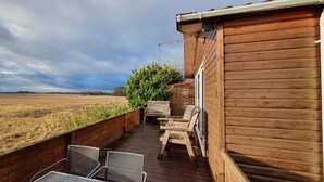 Una terraza de madera con mobiliario exterior, con vistas a un campo y aerogeneradores.